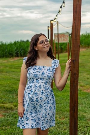 Person in blue floral summer dress poses near wooden post with string lights.