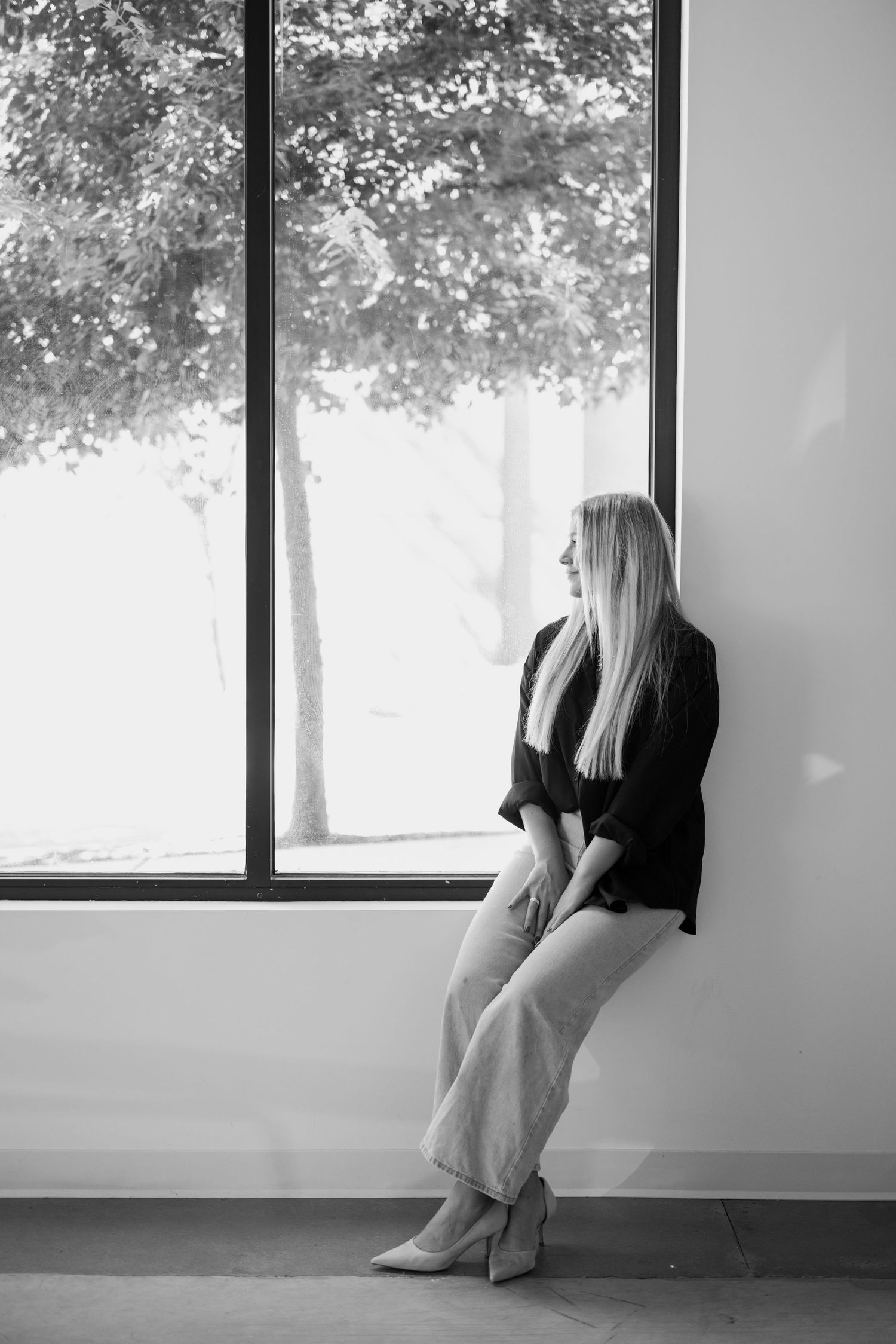 A black and white portrait of someone gazing out a window while leaning against the wall in casual business attire.