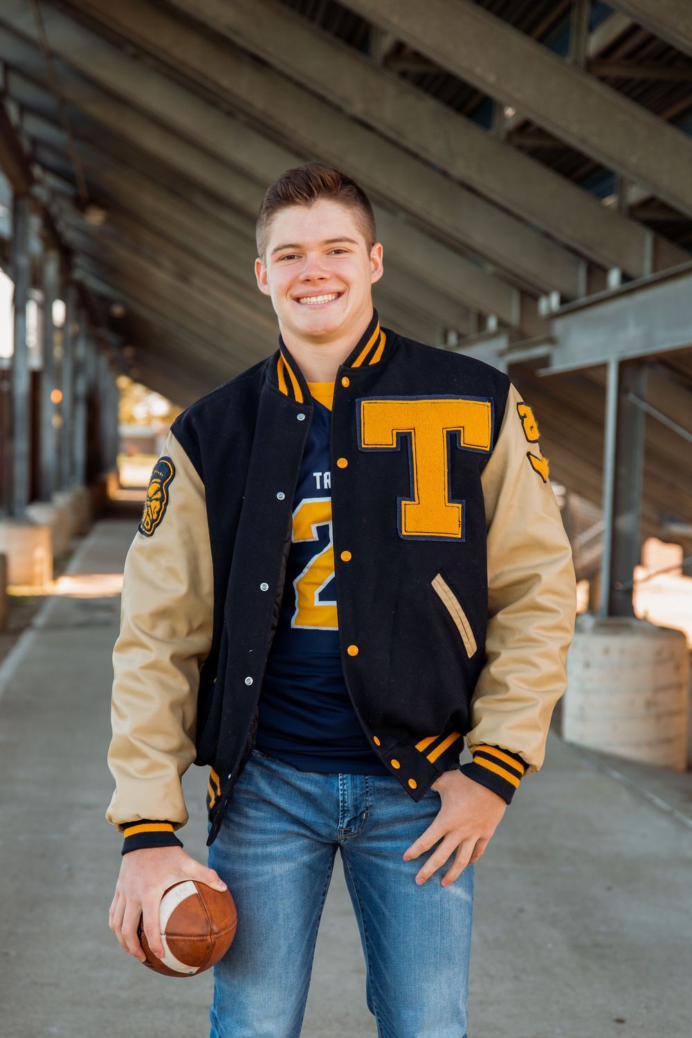 Person wearing varsity letterman jacket holding basketball under concrete structure.