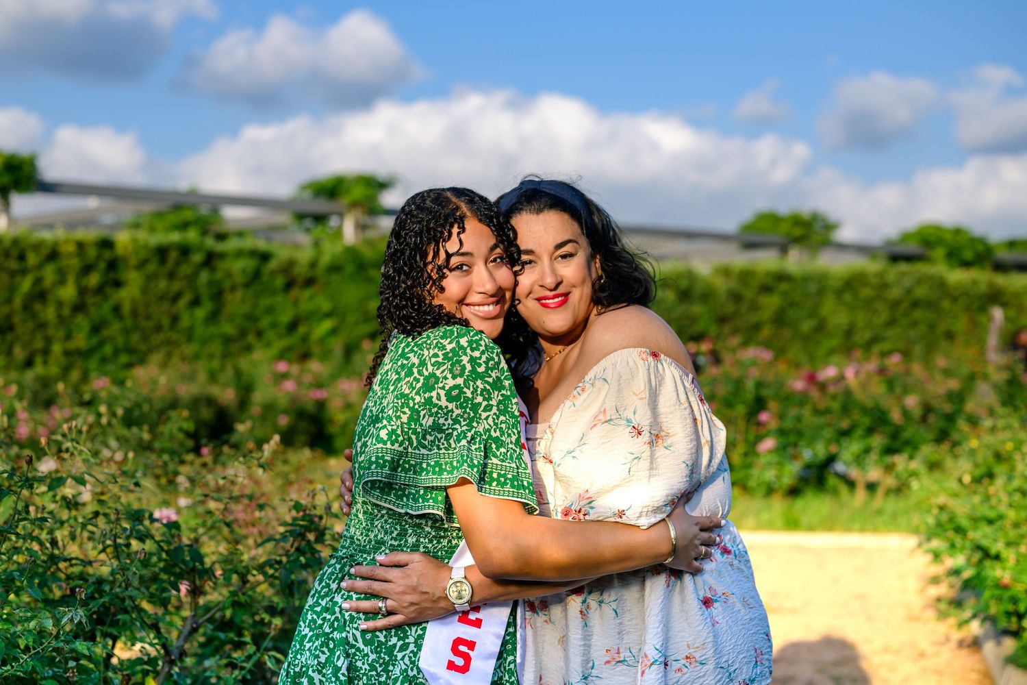 Two people embrace and smile while standing in a sunny field with green foliage and blue skies in the background.