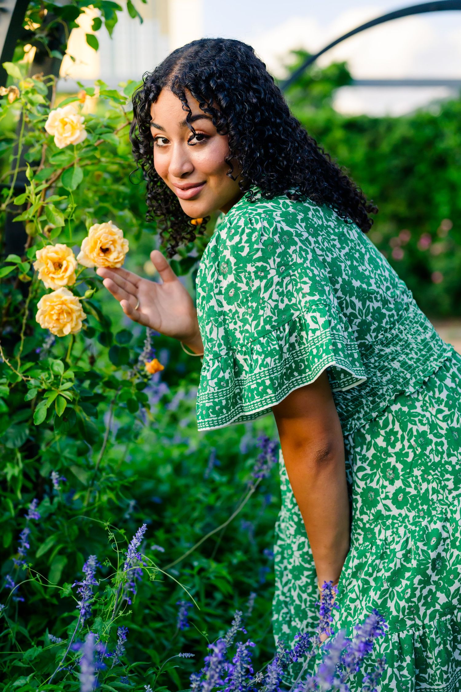 A person in a green dress stands along a garden path beneath a verdant floral archway with a bench nearby.