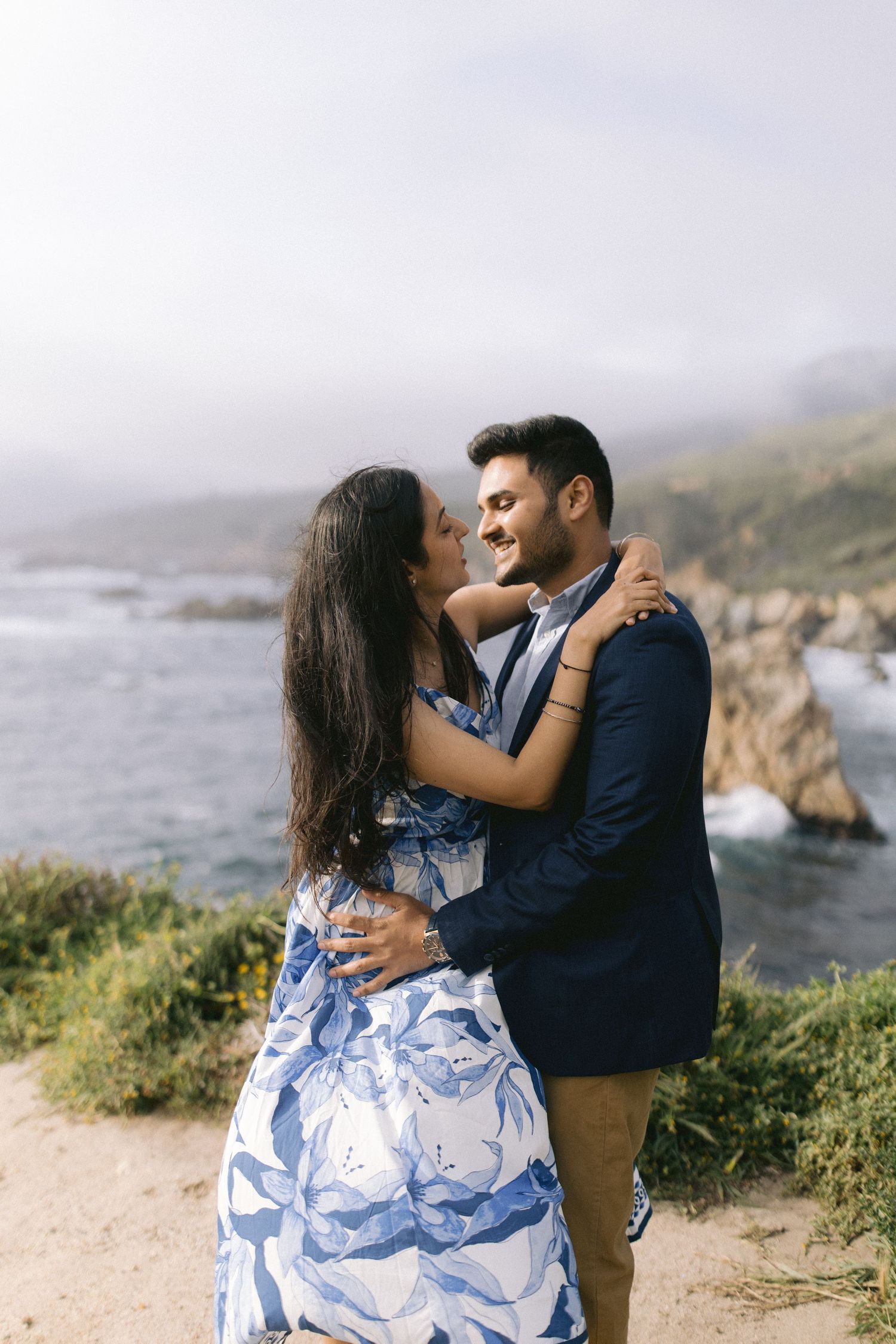 Romantic couple embraces on coastal cliffs overlooking dramatic Big Sur coastline at sunset.