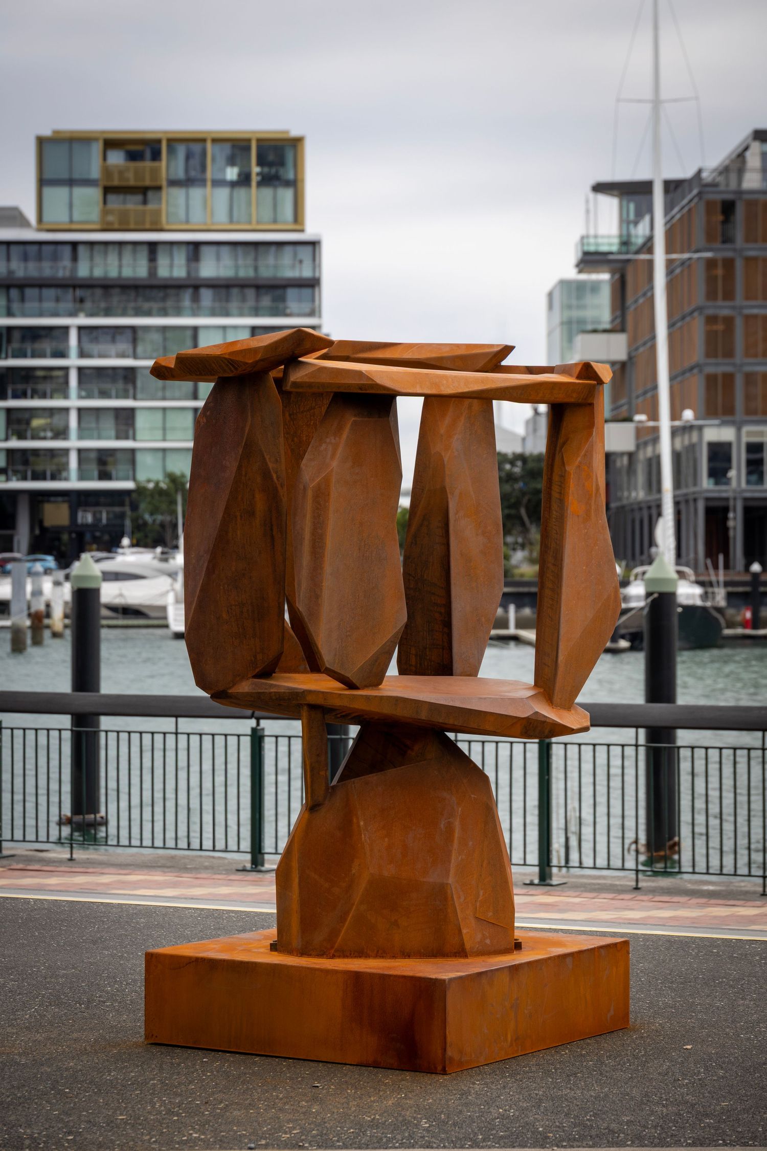 Rust-coloured abstract Corten steel sculpture on a waterfront promenade, with modern buildings and boats in the background.