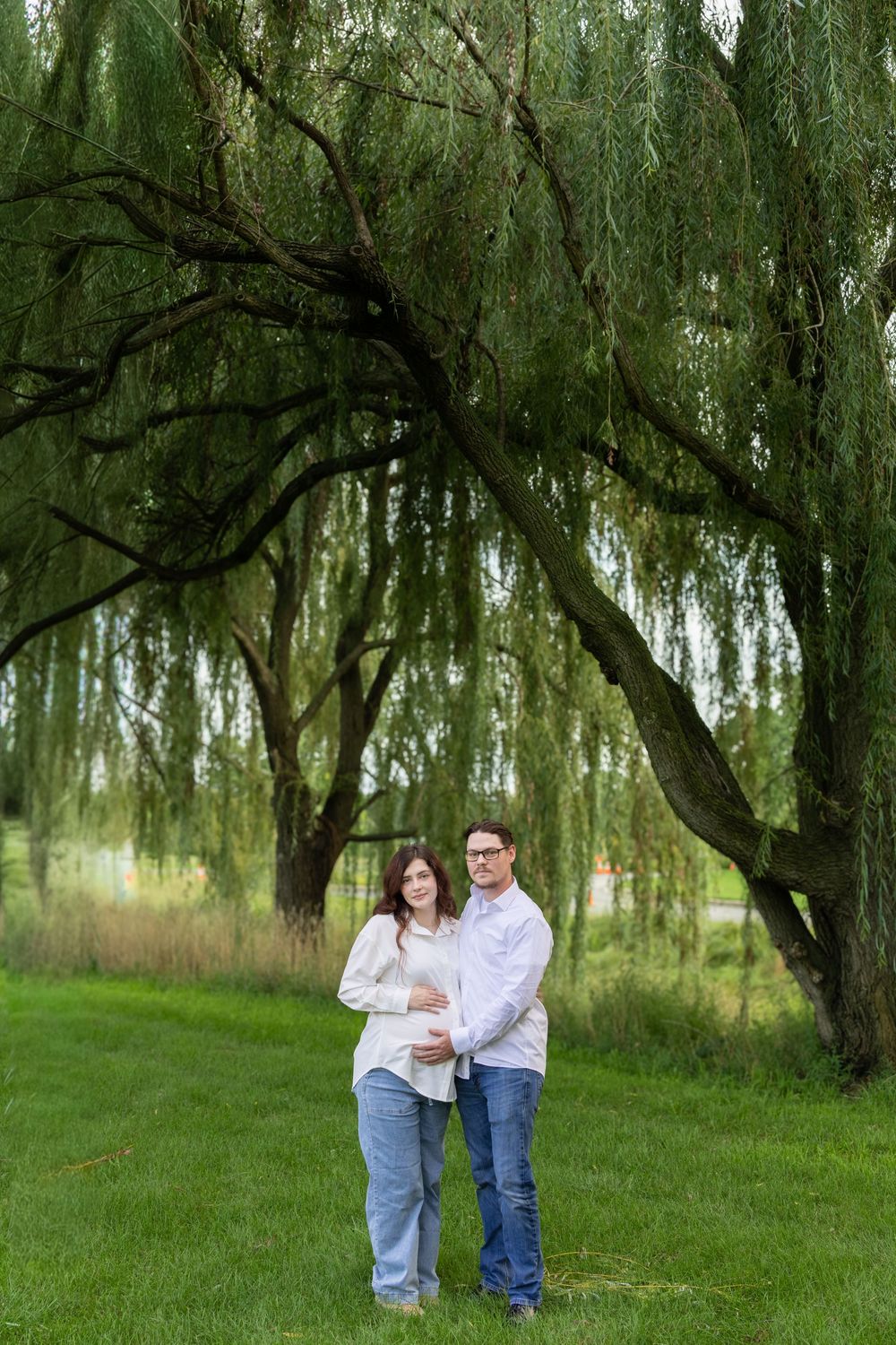 Couple standing under a weeping willow tree on green grass, man embracing pregnant woman in white top and jeans.