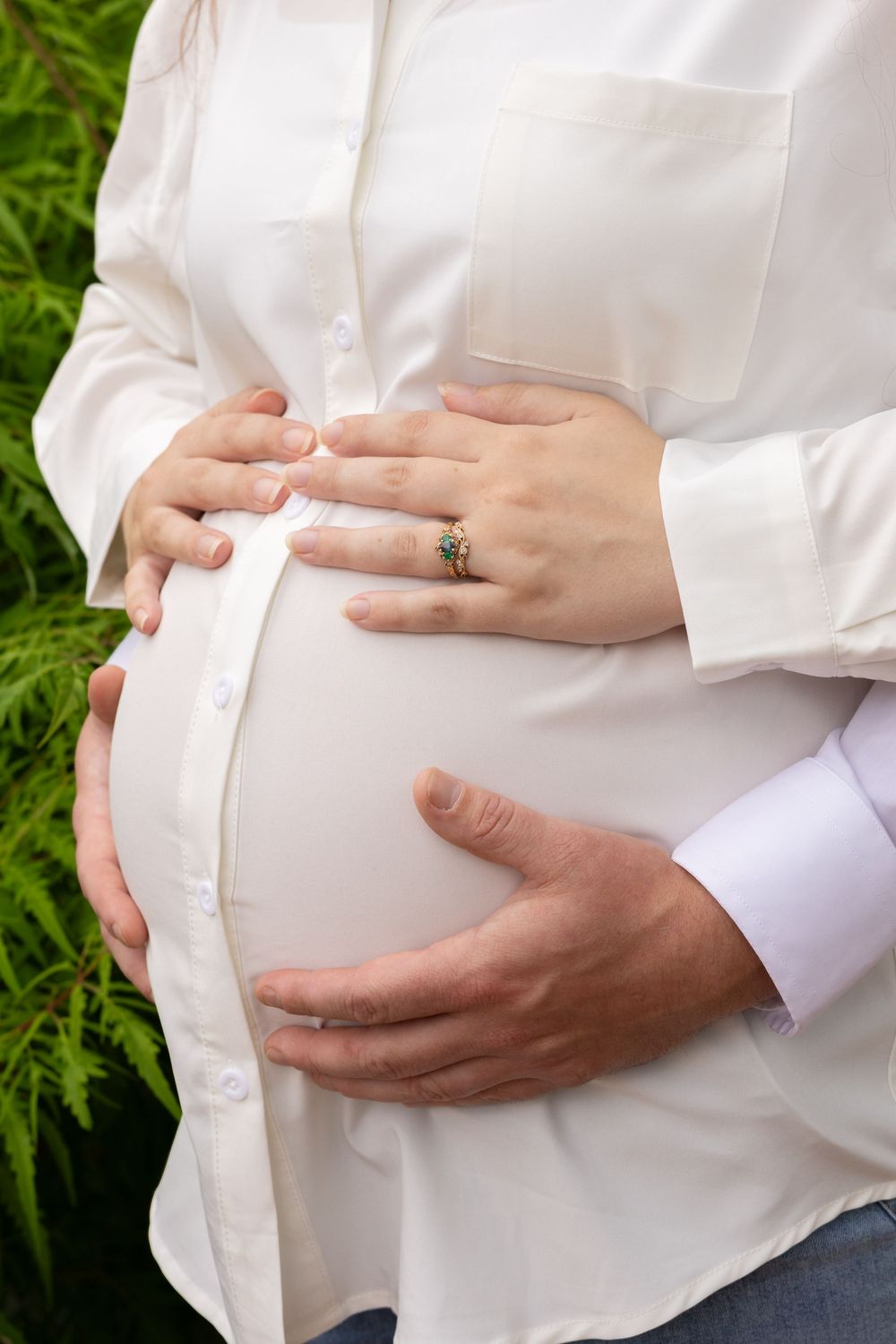 Pregnant woman in white shirt with partner's hands cradling baby bump outdoors near green foliage.