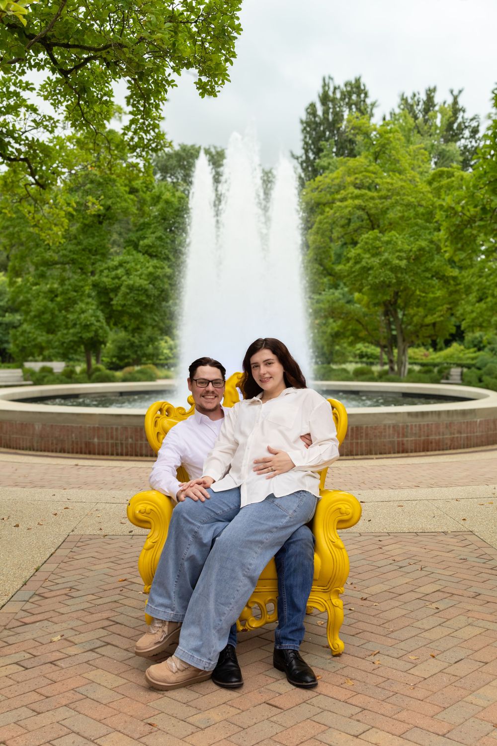 Couple sits on yellow ornate chair in front of fountain in lush green park, woman appears pregnant.