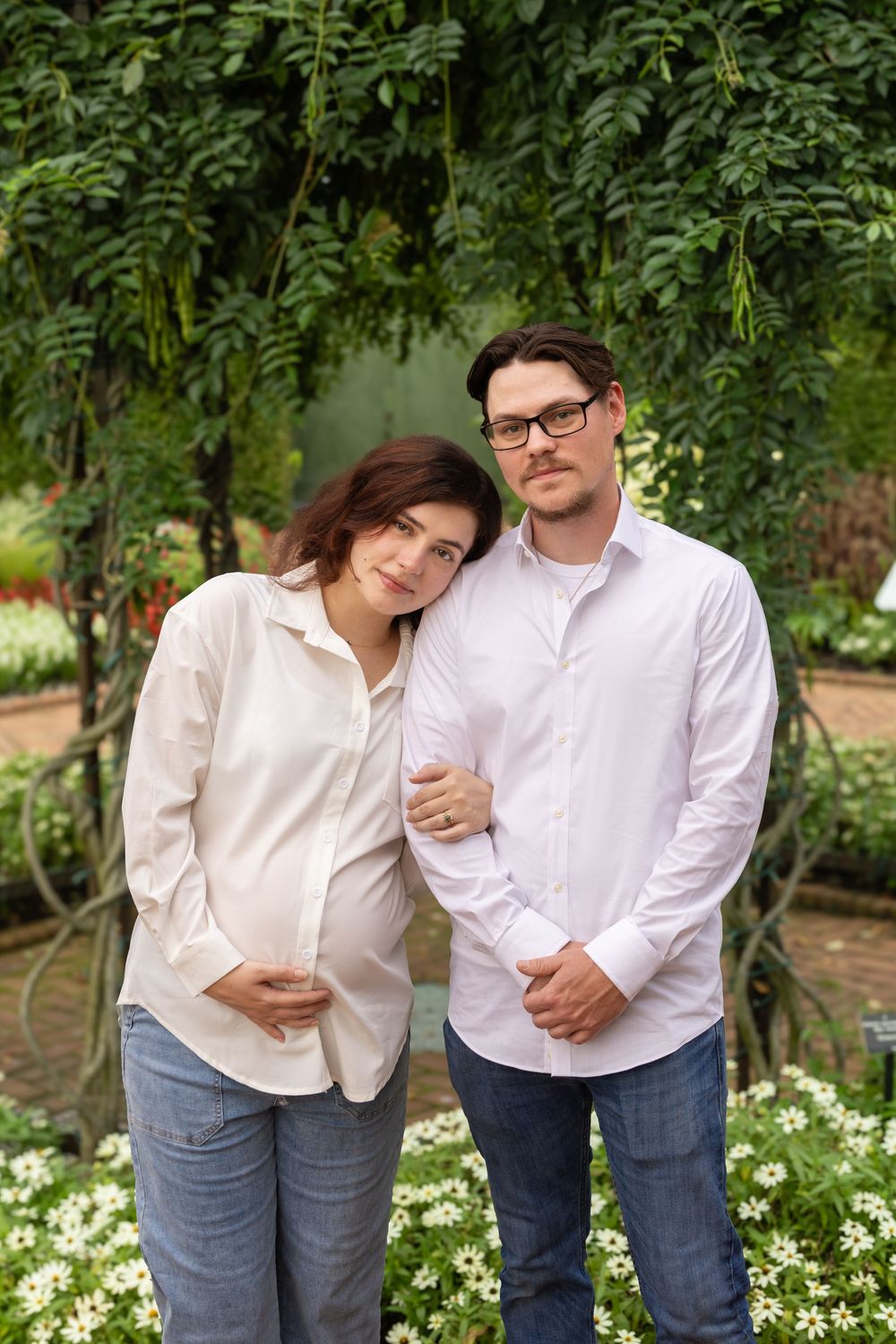 Pregnant woman in white shirt leans on man with glasses outdoors, surrounded by lush greenery and white flowers.