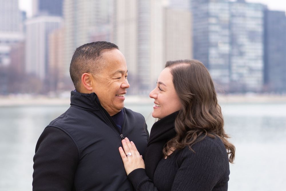 A couple shares a romantic moment together along a waterfront with a city skyline in the background.