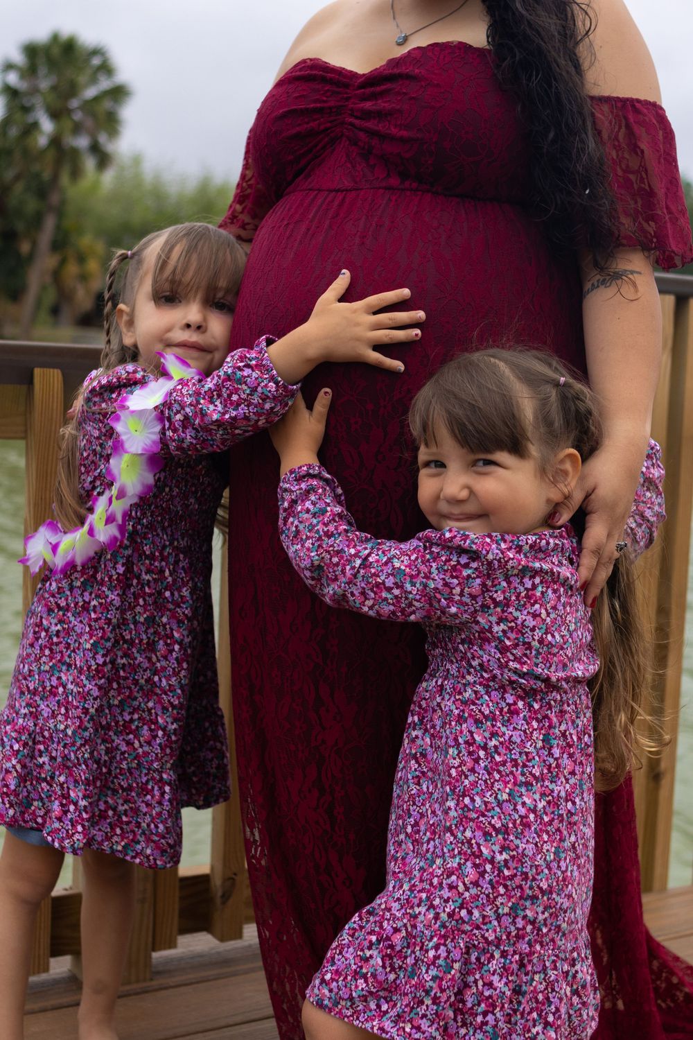 Two young girls in floral dresses hugging their pregnant mother in a red maternity gown outdoors.