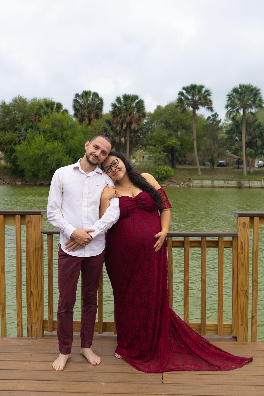 Couple posing on wooden dock, pregnant woman in red gown leaning on partner, lake and palm trees background.
