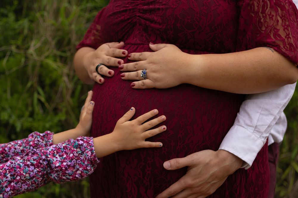 Close-up of pregnant woman in red lace dress with partner and child's hands gently touching her baby bump outdoors.