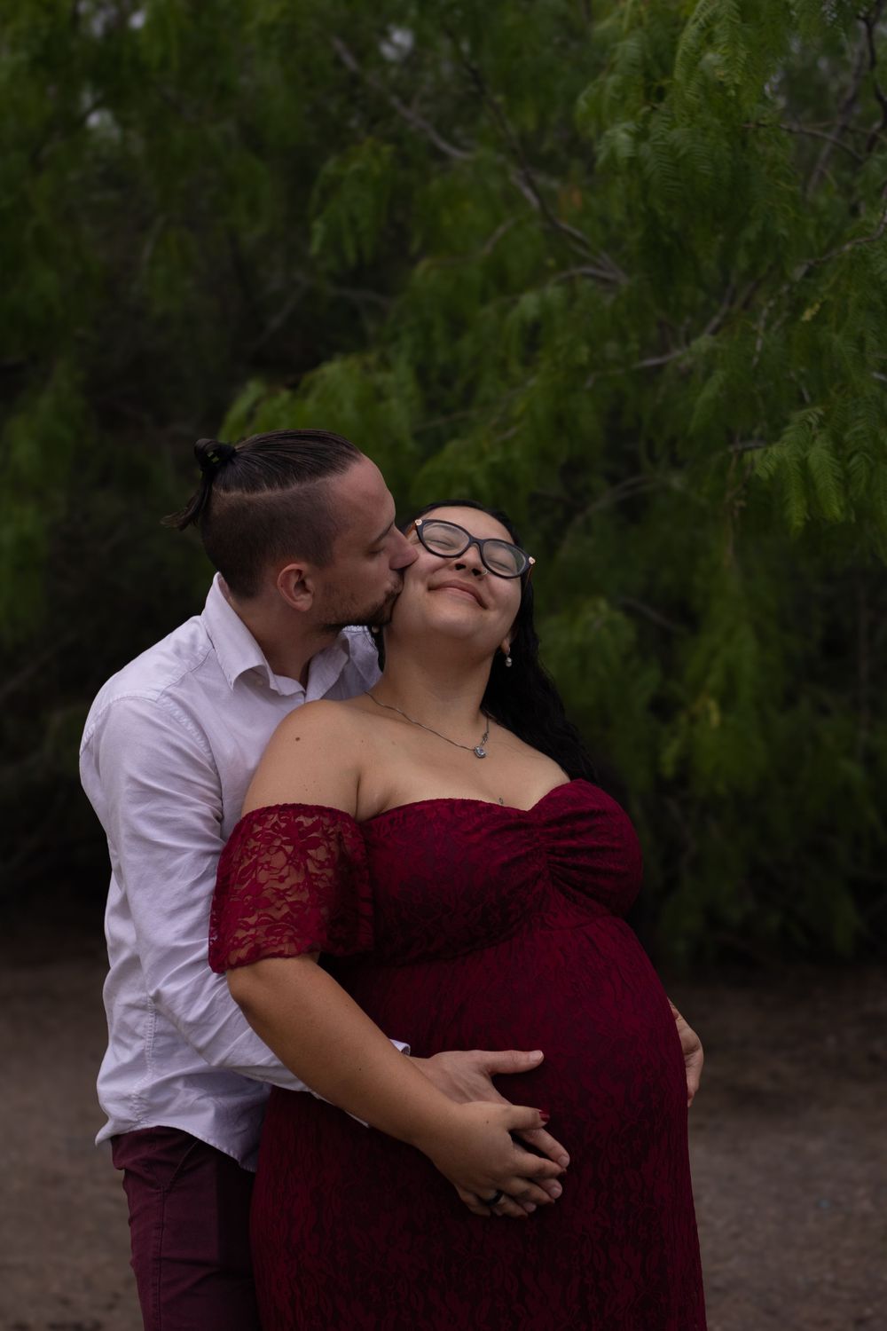 Couple sharing a tender kiss outdoors, man in white shirt embracing pregnant woman in elegant red lace maternity dress.