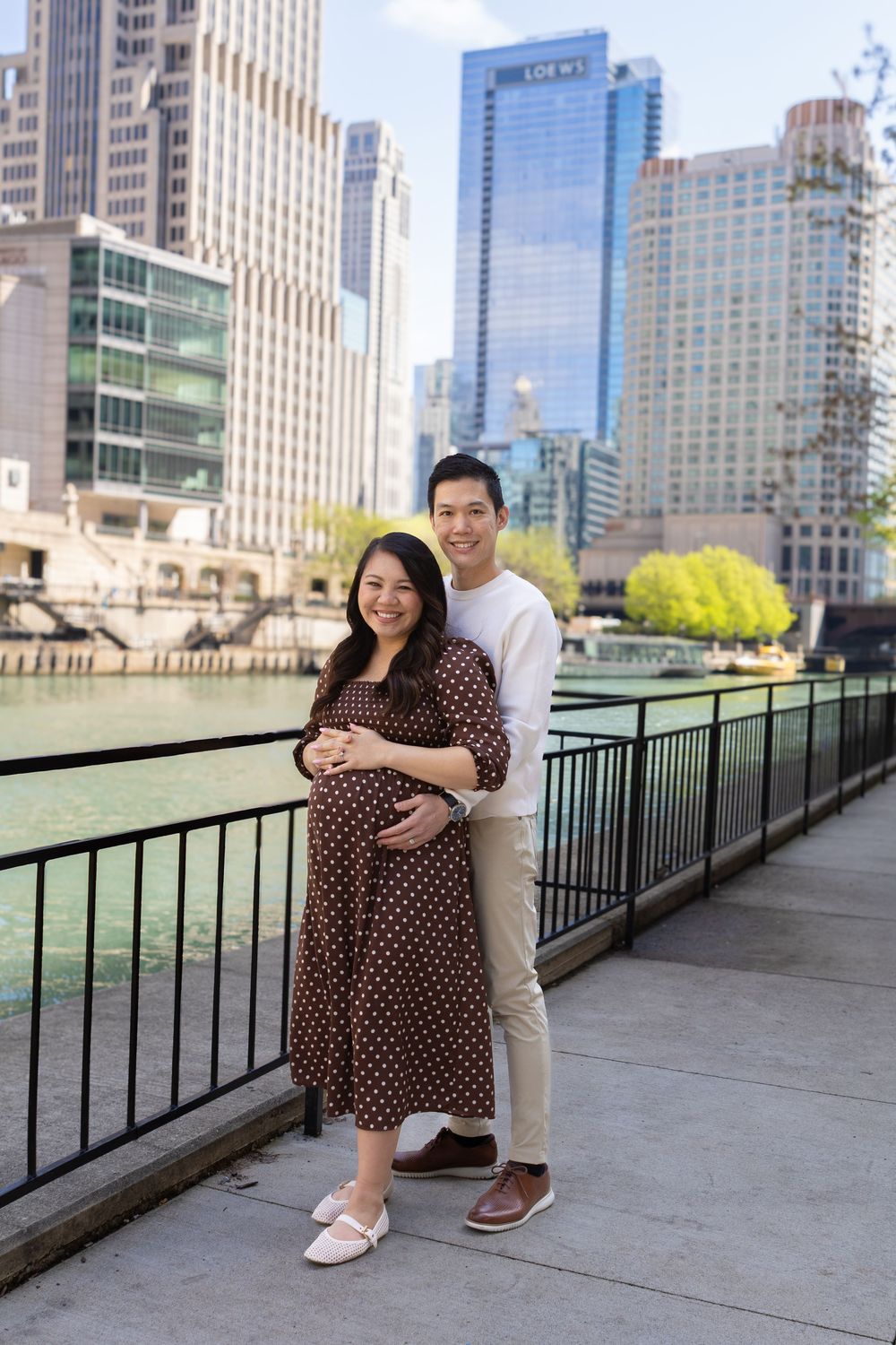 Pregnant couple posing on Chicago Riverwalk with city skyline, woman in polka dot dress, man in white sweater.