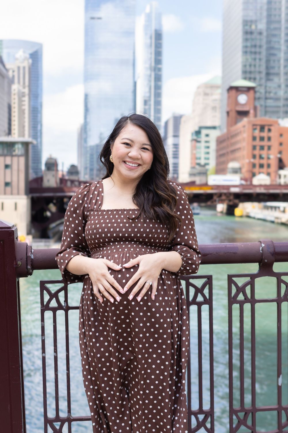 Smiling pregnant woman in brown polka dot dress posing on Chicago Riverwalk bridge with city skyline backdrop.