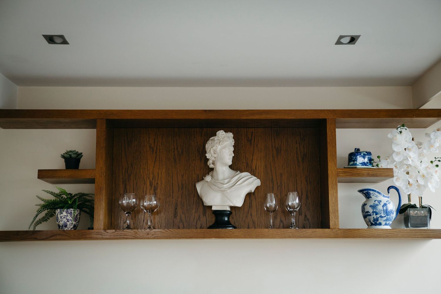 A white classical bust sculpture sits on wooden shelving alongside blue and white porcelain vases and crystal glassware.