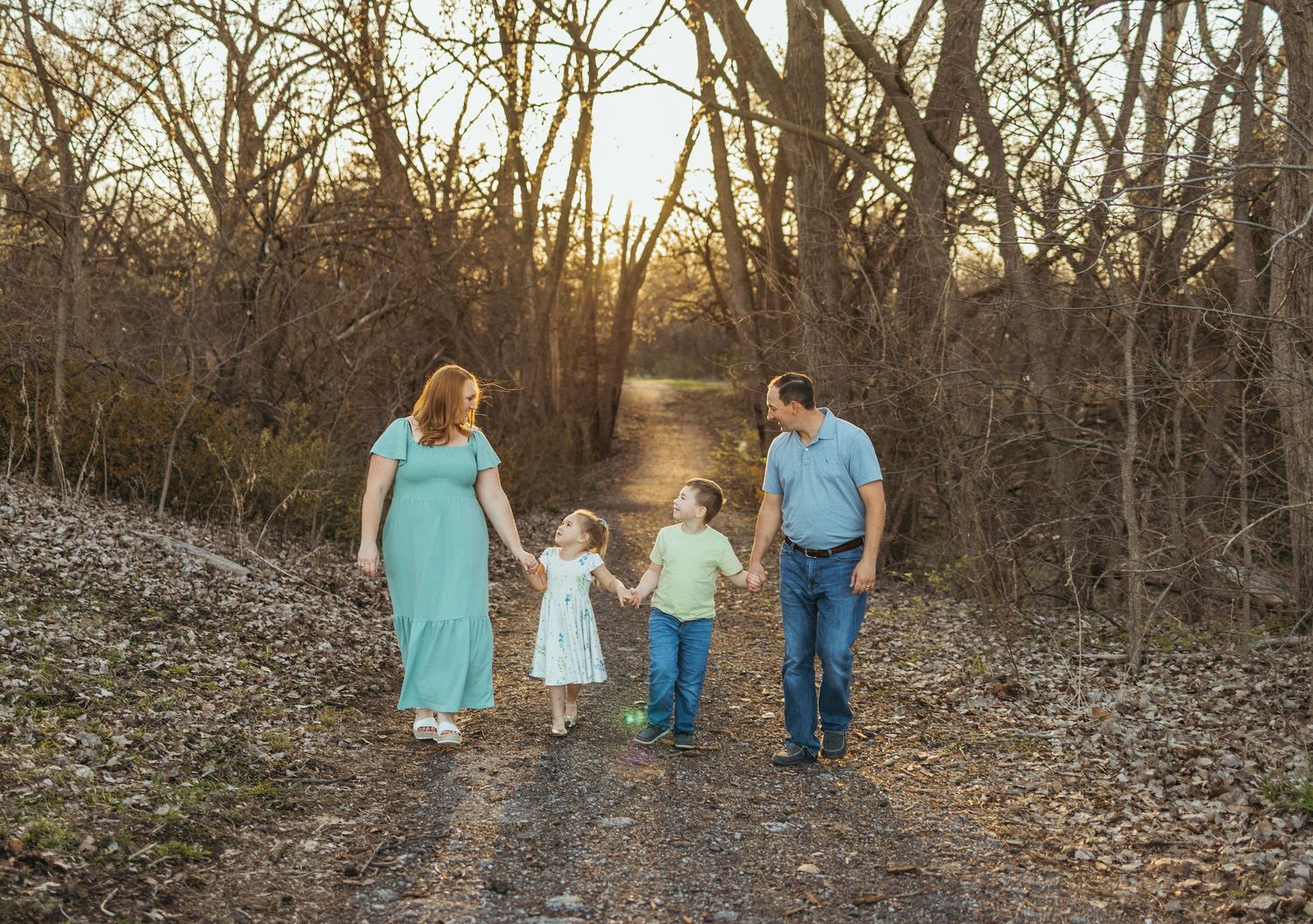 A family in coordinating blue and white outfits walks hand in hand along a wooded path during sunset.