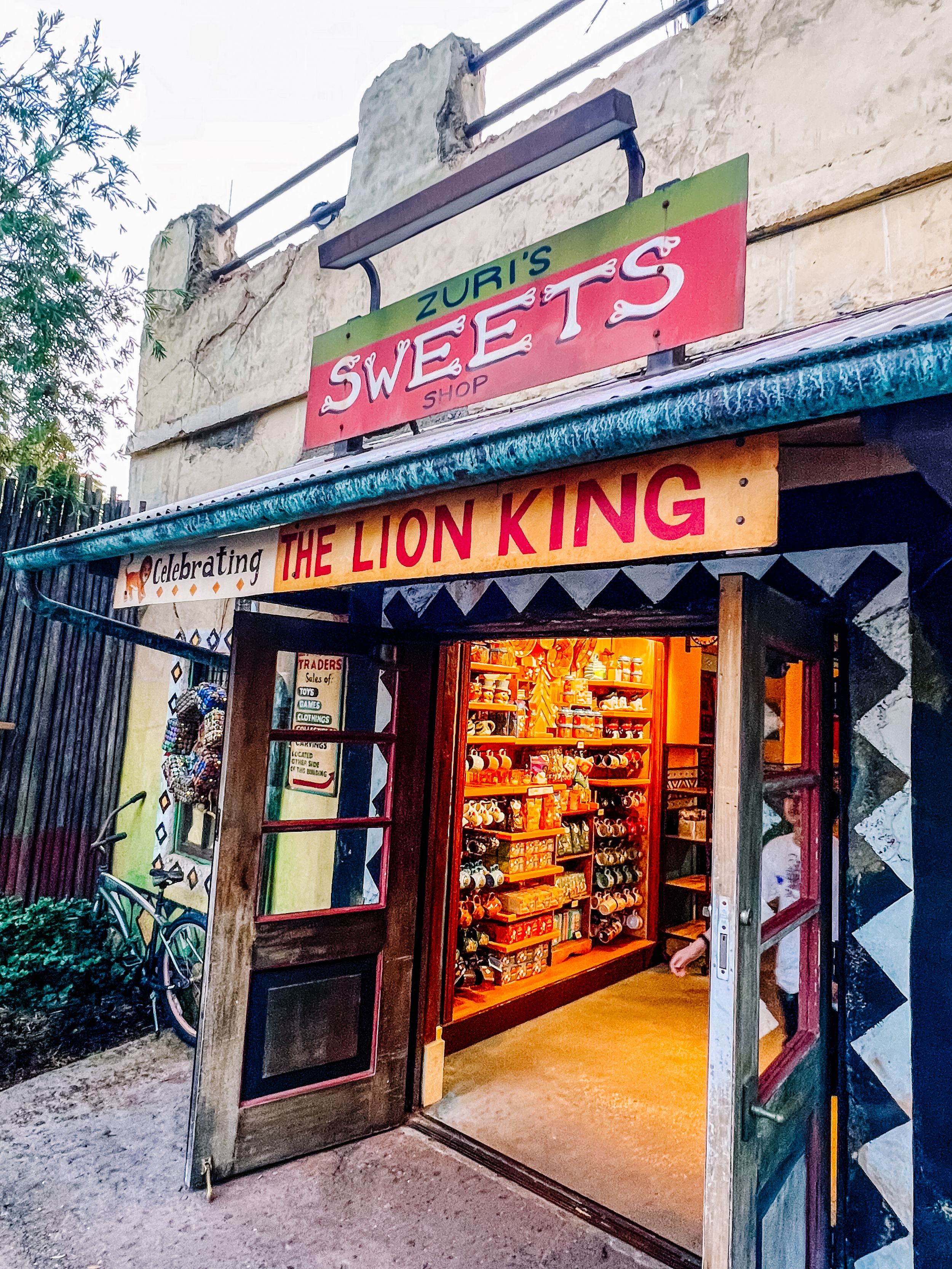 Storefront of Zuri's Sweets shop featuring The Lion King merchandise display with glowing interior visible through open door.