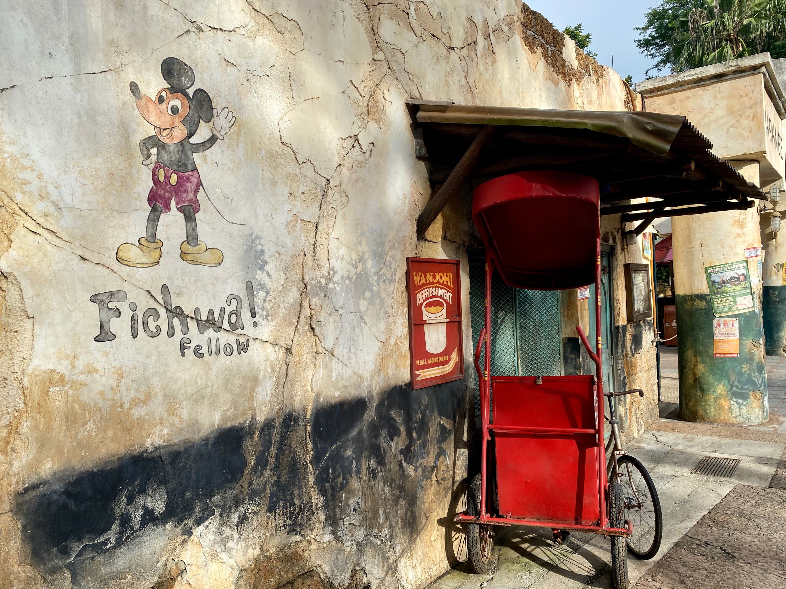 A vintage Mickey Mouse mural painted on a weathered wall next to a red cart stand in a tropical street setting.