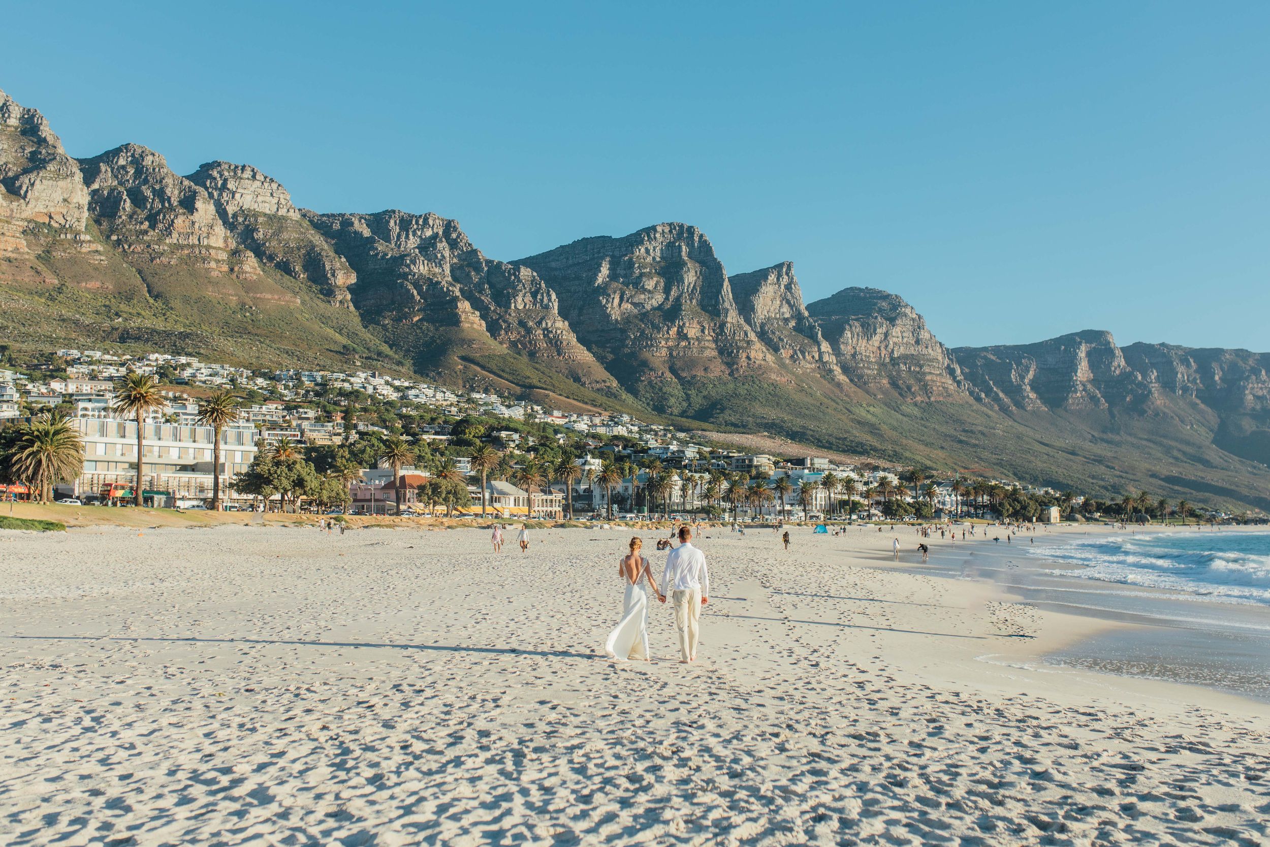 Two figures walking on Camps Bay beach with the Twelve Apostles mountain range towering in background on a clear day in Cape Town.