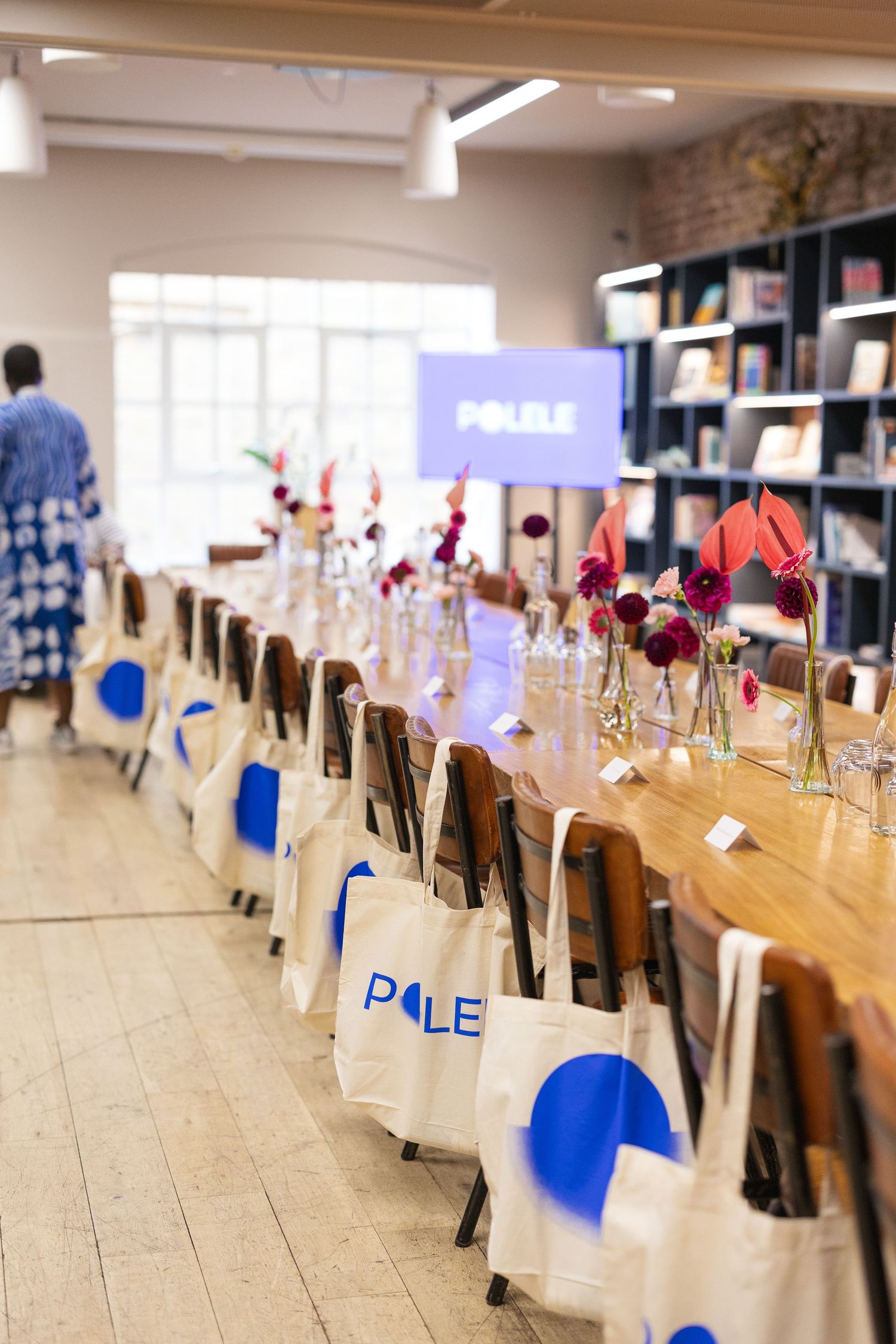 Long wooden table decorated with blue and white tote bags and red flowers in vases for a special event setup.