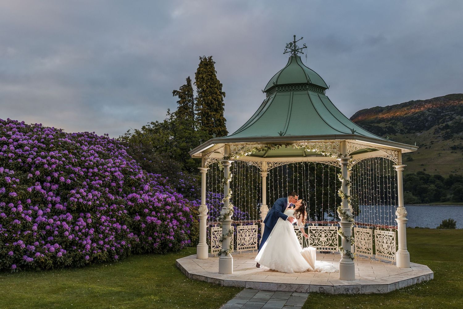 A scenic wedding venue with a green-roofed gazebo and blooming purple rhododendrons overlooking a serene lake.