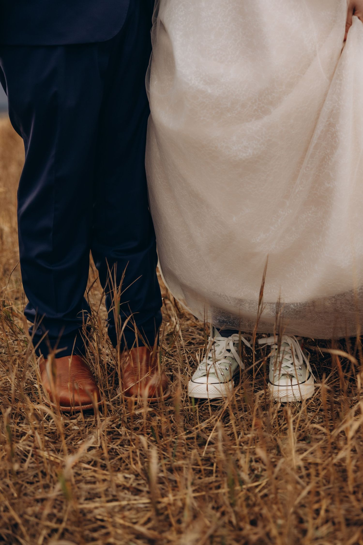 Close-up shot of wedding couple's feet with white sneakers and dress shoes standing on dried grass.