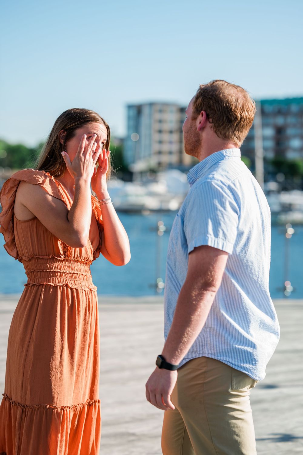 A couple shares a joyful moment on a waterfront boardwalk with boats and buildings in the background.