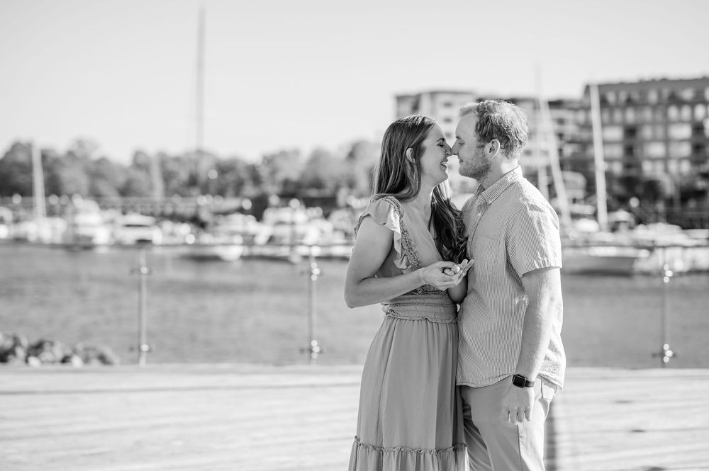 A couple  shares an intimate moment by a marina with boats and buildings in the background. Harbor Point Boardwalk in Stamford, CT