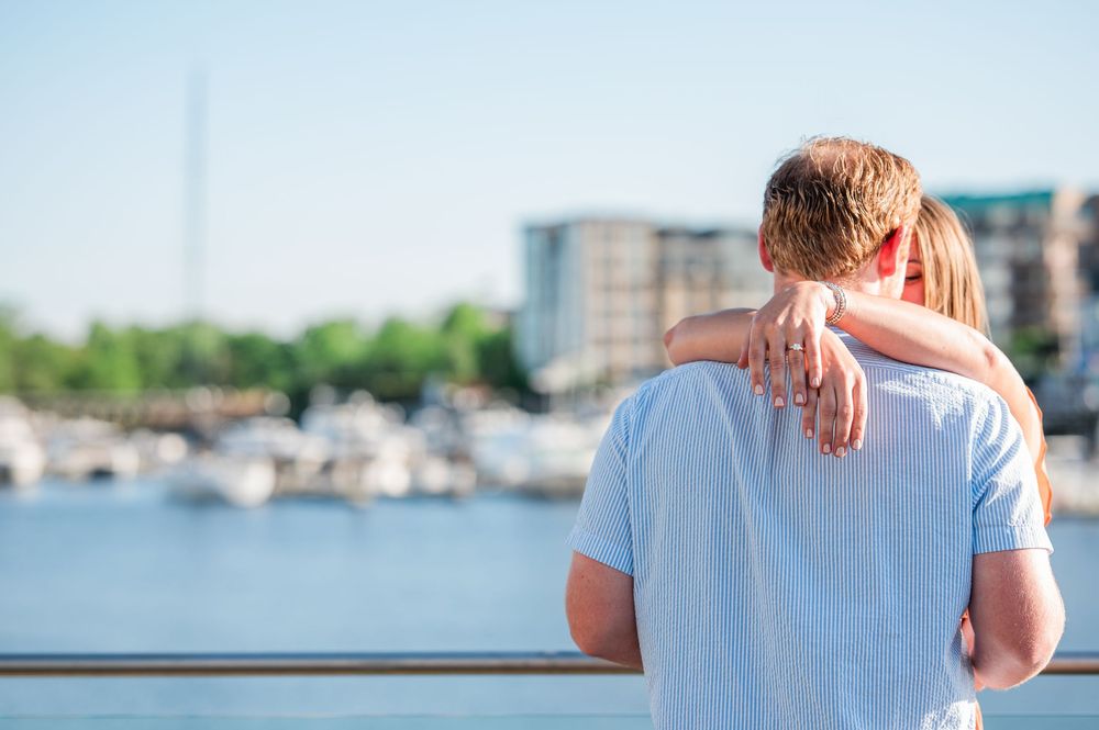 A person in a white t-shirt holds a child while looking out over a marina with boats and buildings in the background.