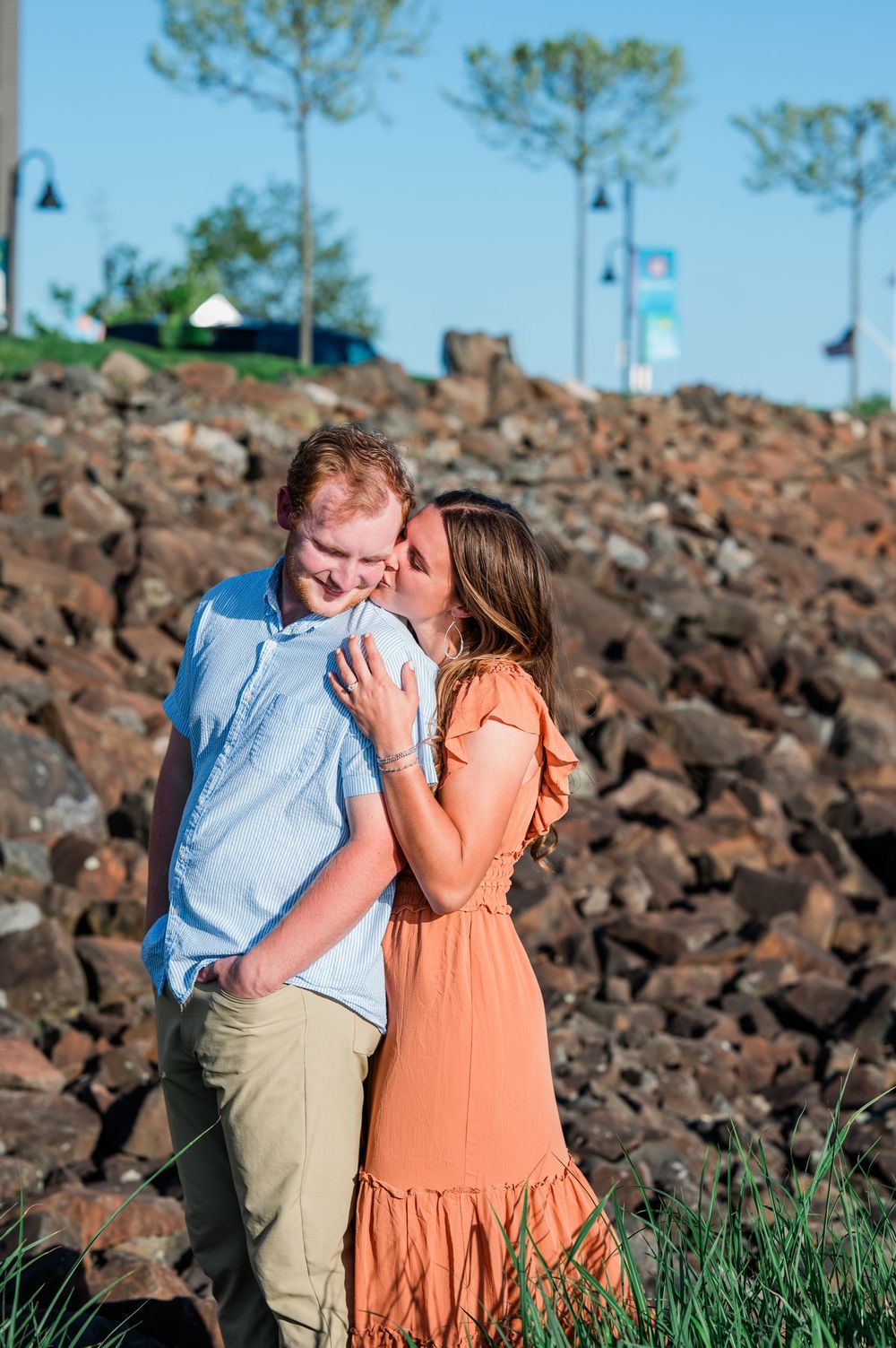 Two people embrace and laugh together among rocky terrain and grass near the waterfront. Harbor Point Boardwalk in Stamford, CT