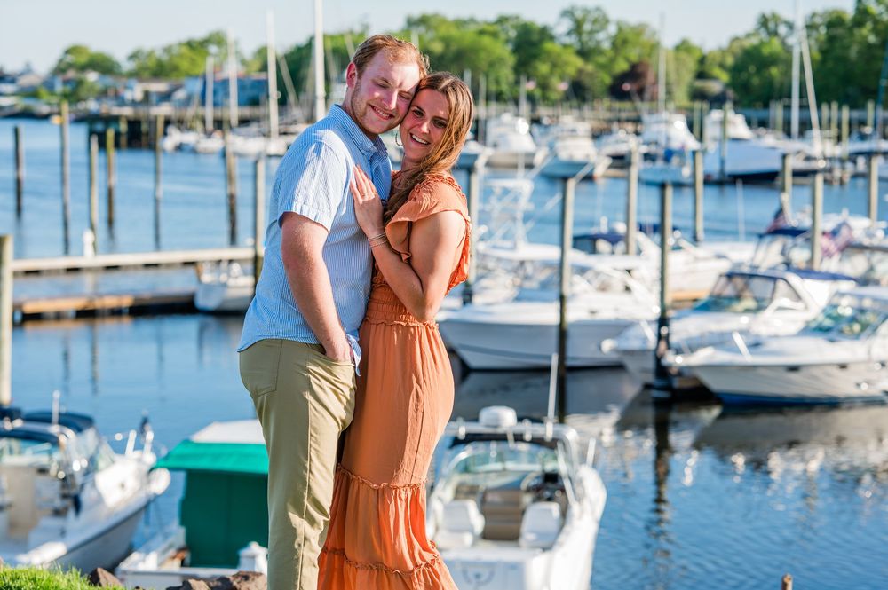 A couple embraces by a marina filled with boats on a beautiful summer day. Harbor Point Boardwalk in Stamford, CT