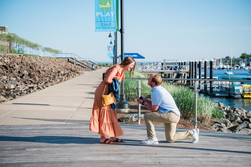 Romantic couple embraces on a sunny boardwalk by a marina with boats and blue sky in the background. Harbor Point Boardwalk in Stamford, CT