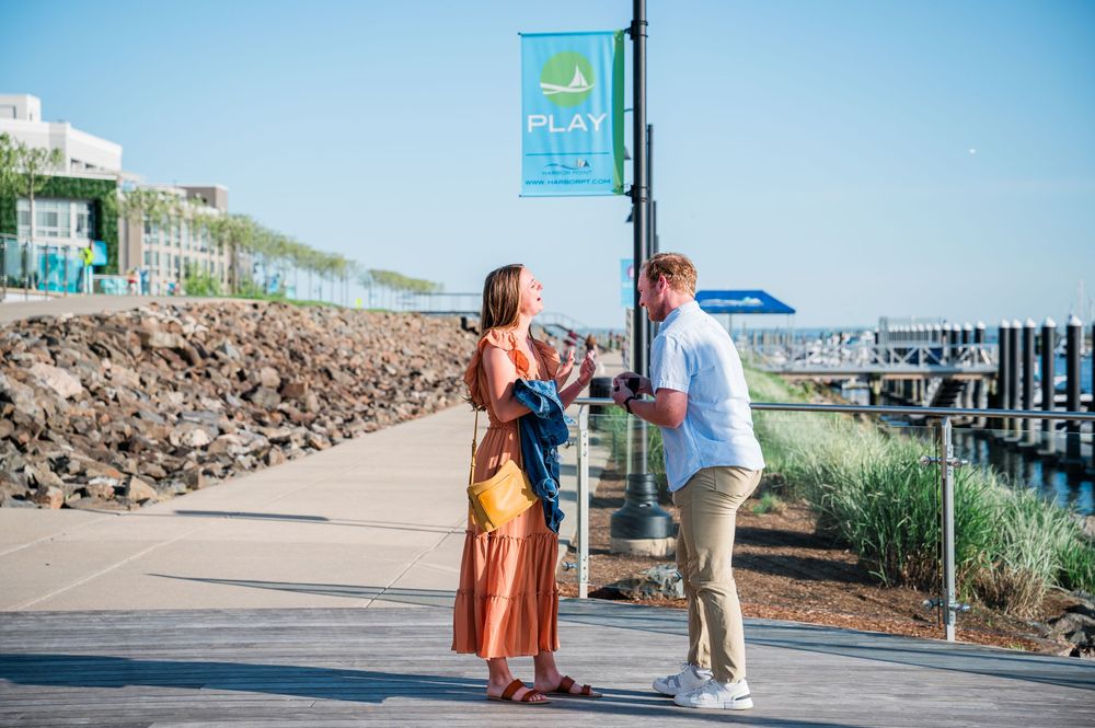 A couple stands together on a coastal walkway with a harbor view on a sunny summer day.