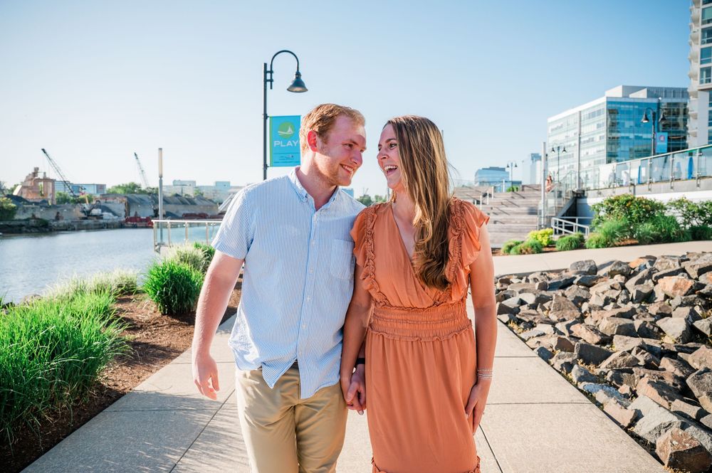A romantic moment shared between a couple on a waterfront path with boats and buildings visible. Harbor Point Boardwalk in Stamford, CT