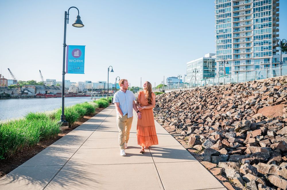A couple walks along a waterfront path with city buildings and rocky shoreline in background. Harbor Point Boardwalk in Stamford, CT