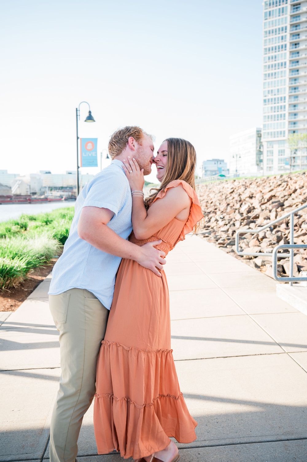 Couple shares an intimate moment on a waterfront walkway with high-rise buildings in the background. Harbor Point Boardwalk in Stamford, CT