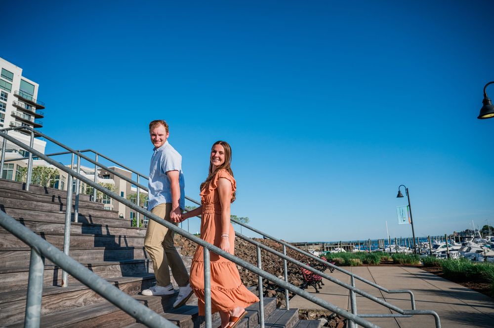 A couple descends outdoor stairs together against a bright blue sky and harbor backdrop. Harbor Point Boardwalk in Stamford, CT