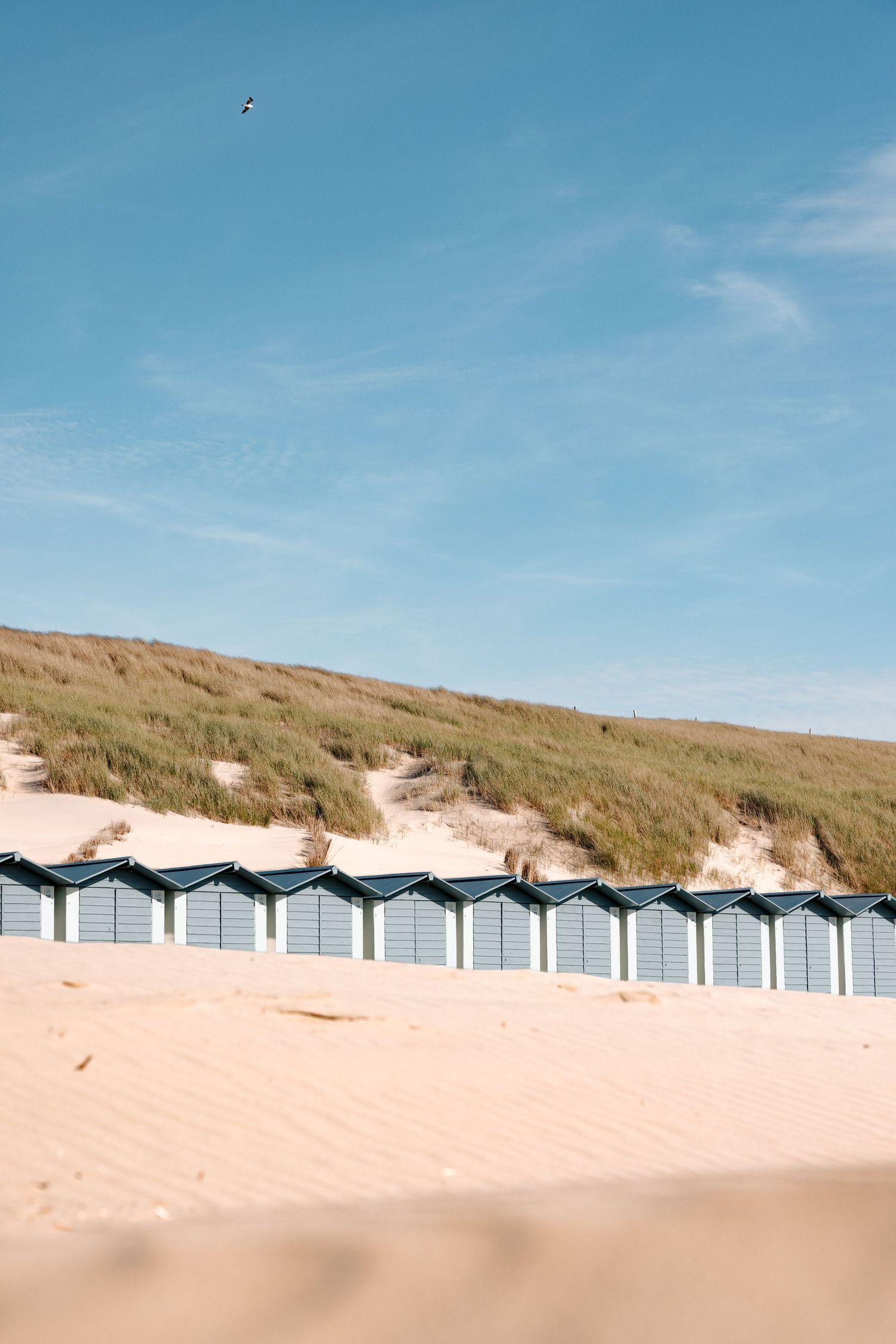 “Een bruidspaar maakt een strandwandeling op blote voeten langs strandhokjes aan de Noord-Hollandse kust tijdens een trouwshoot.”
