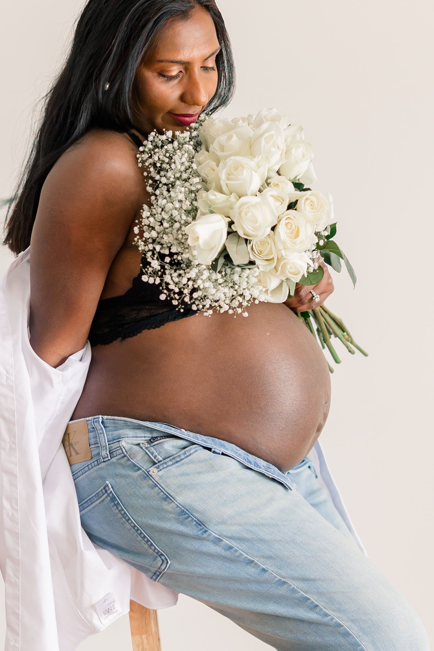 A person in light denim jeans holds a delicate bouquet of white roses and baby's breath against a white background.
