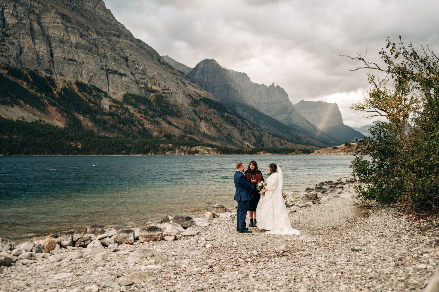A panoramic view of a mountain lake wedding ceremony set against dramatic rocky peaks and cloudy skies.