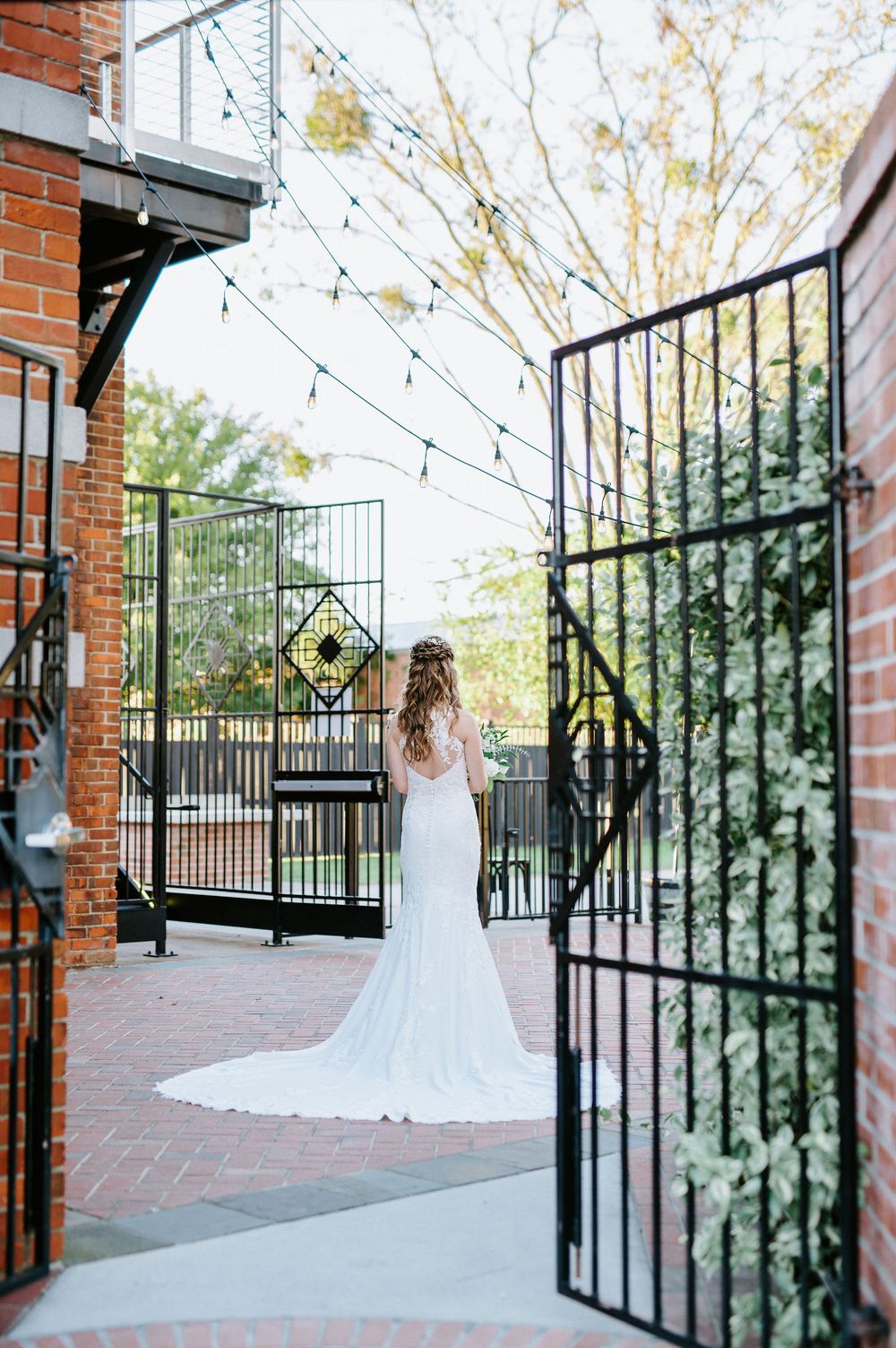 A white wedding dress flows elegantly against brick walls and wrought iron gates in an outdoor courtyard setting.