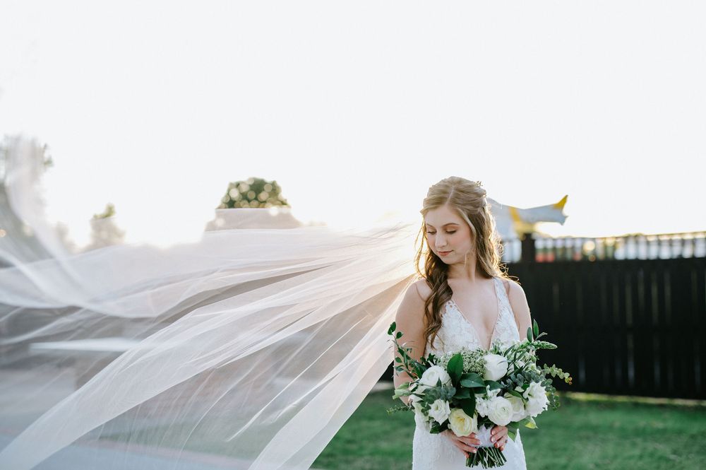 A flowing white veil catches the breeze as it frames a romantic bridal portrait with lush greenery and flowers.