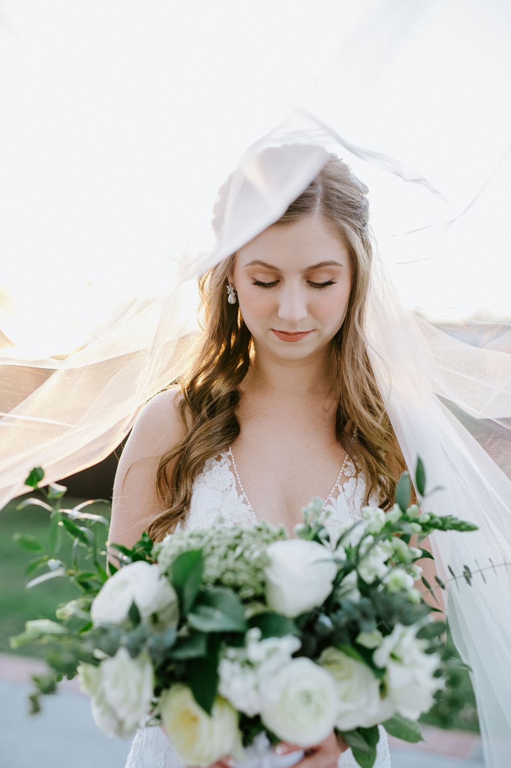 An elegant white bridal bouquet with roses and greenery is held against a delicate lace wedding gown.