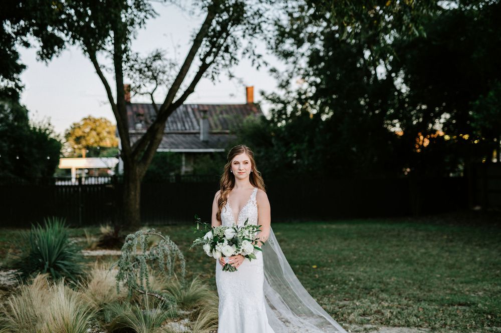 A bride in a white lace gown holds a colorful bouquet while standing in a garden setting at dusk.