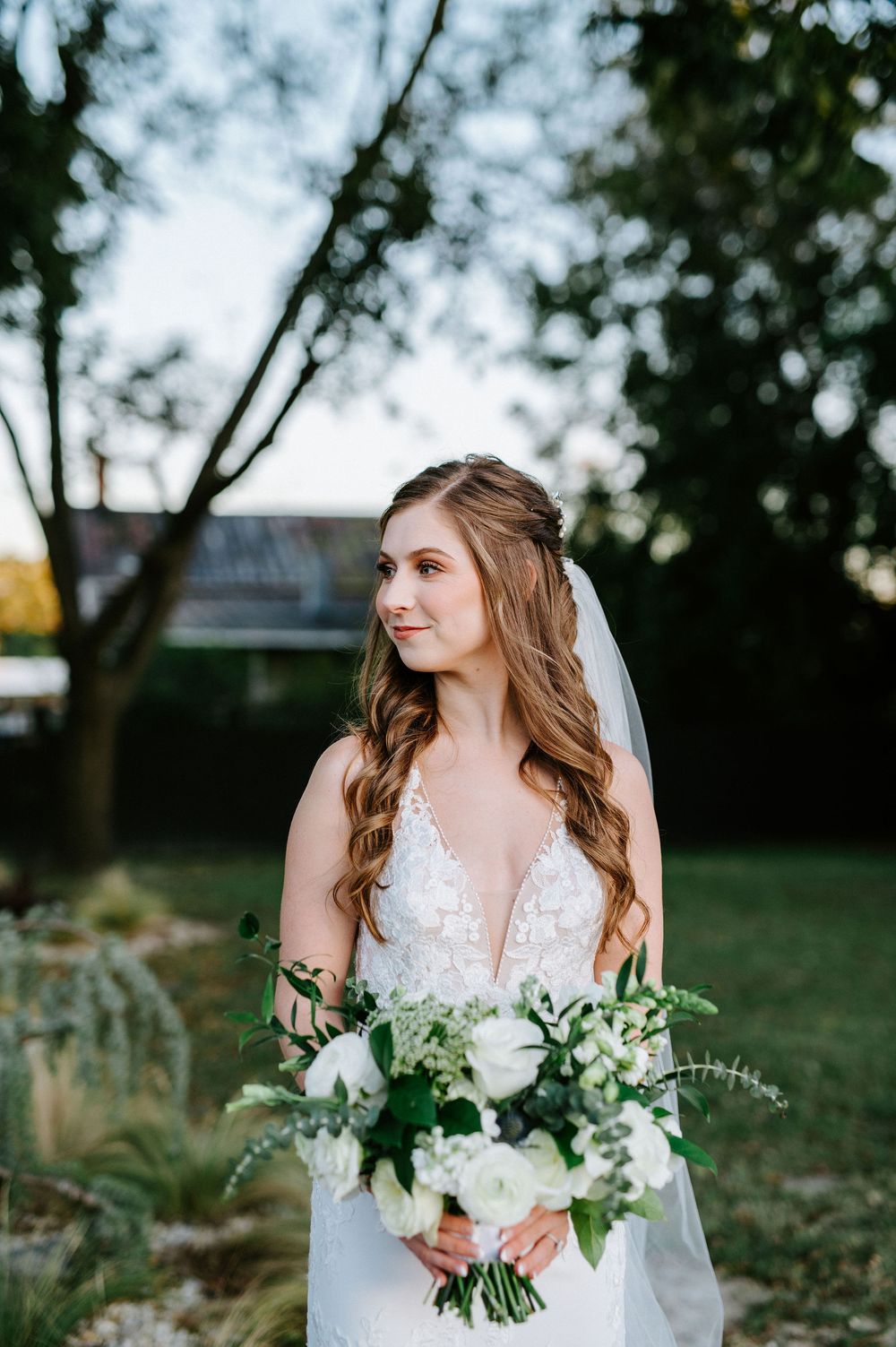 Bride in lace wedding dress holds white floral bouquet with greenery while standing in outdoor garden setting.