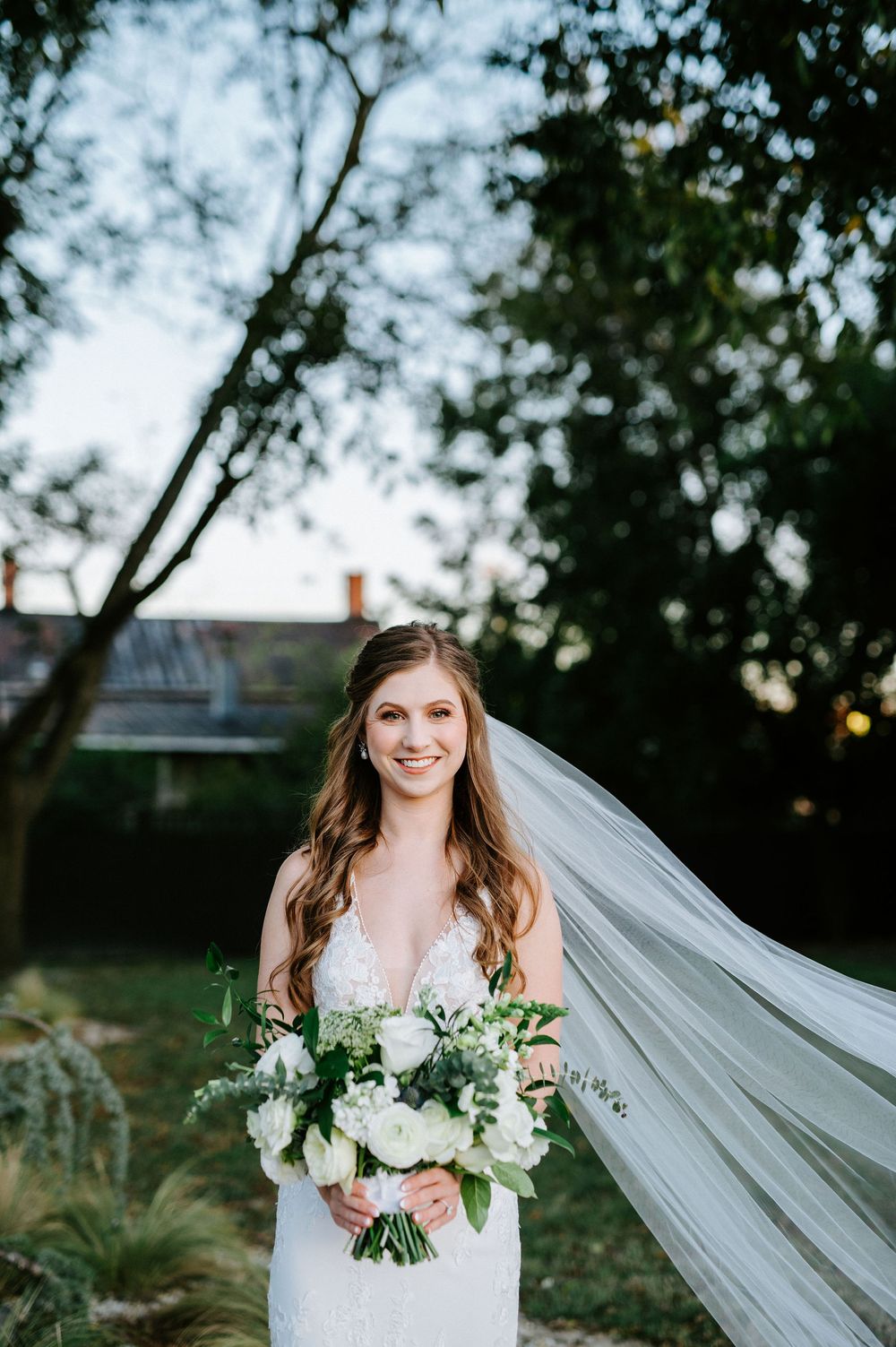 Bride in white lace dress with flowing veil and white floral bouquet poses outdoors at dusk.