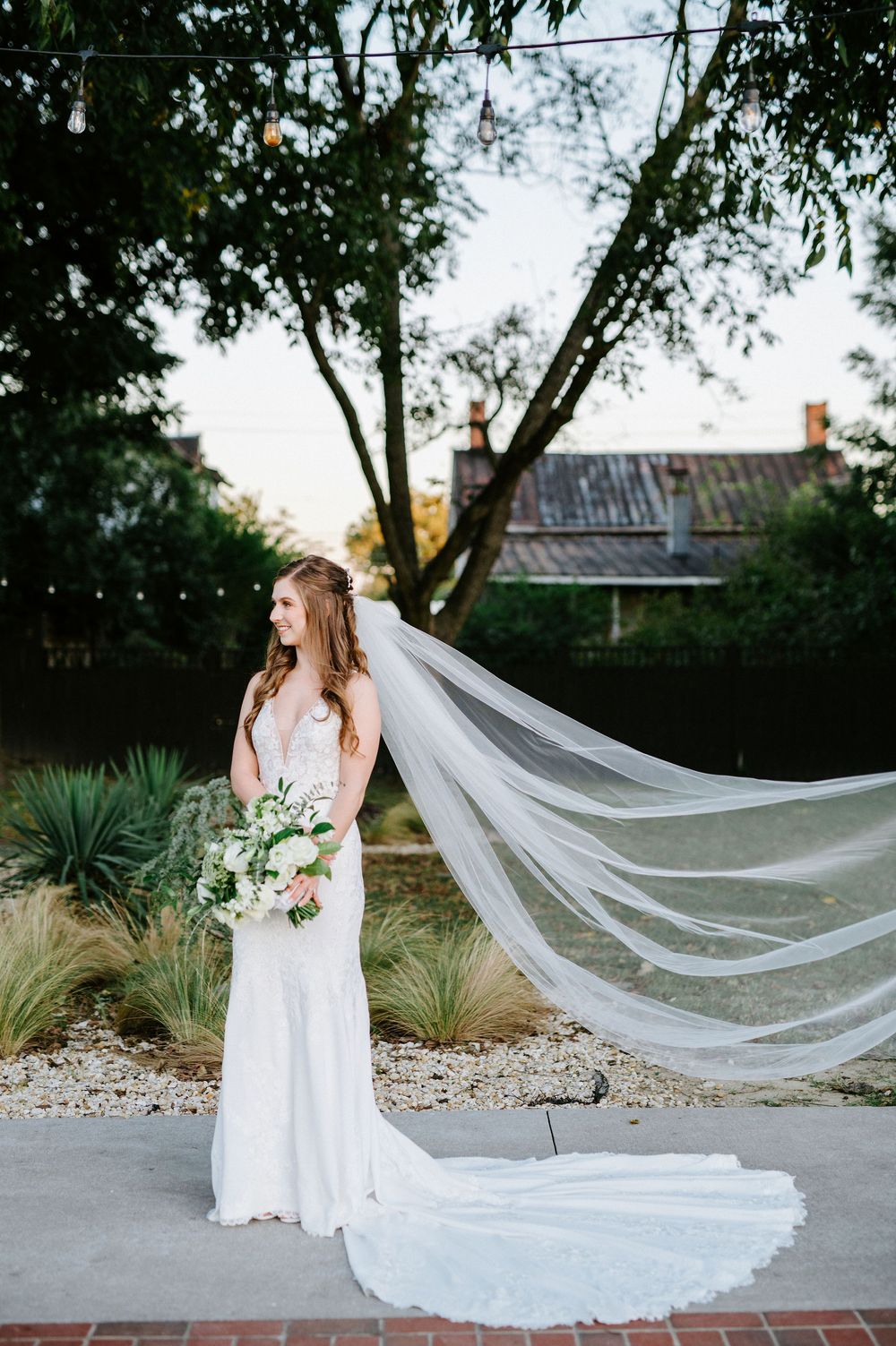A flowing cathedral length veil trails behind a bride in a white lace gown standing in a garden setting.