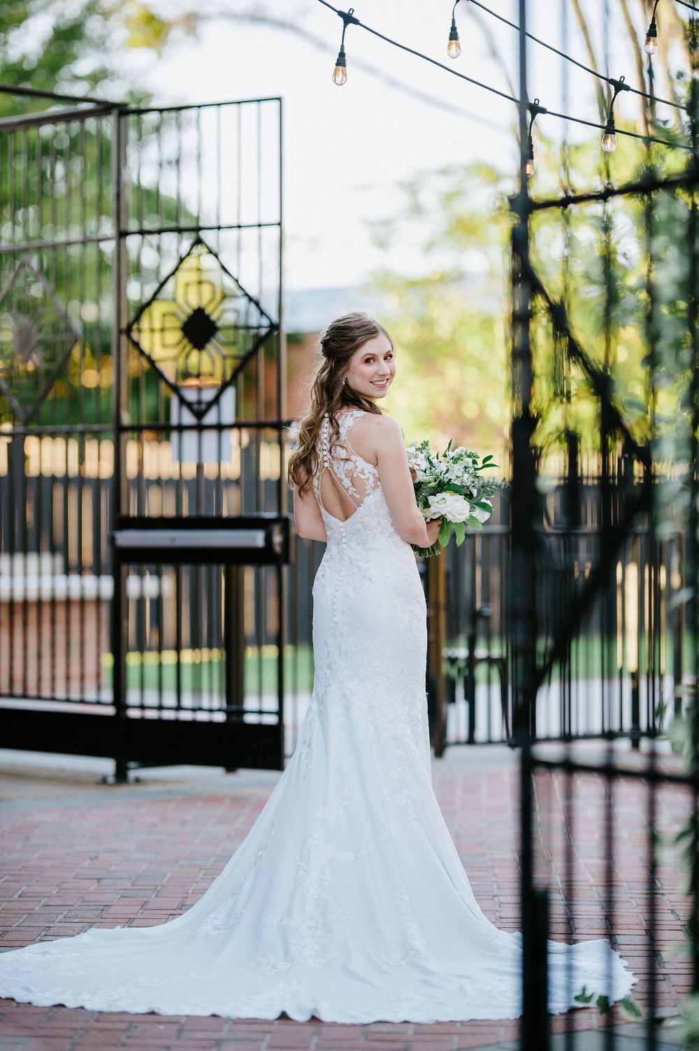 A bride in a white lace wedding dress poses near a black iron gate in an outdoor garden setting.