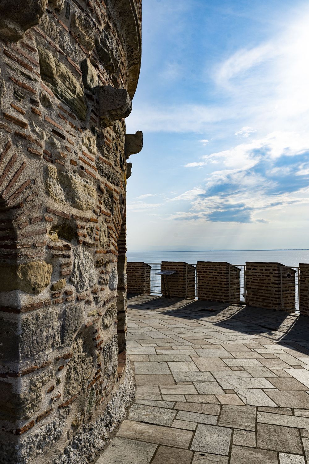 Stone battlements of a medieval castle wall overlook a serene ocean horizon under a bright blue sky with scattered clouds.