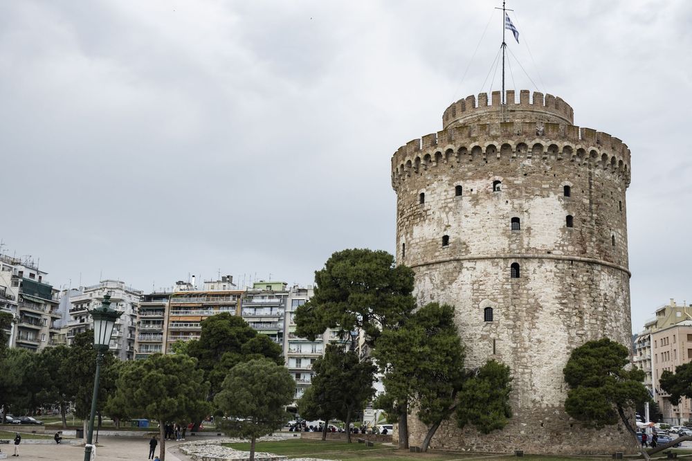 The iconic White Tower of Thessaloniki stands tall against a cloudy sky in Greece with modern buildings in the background.